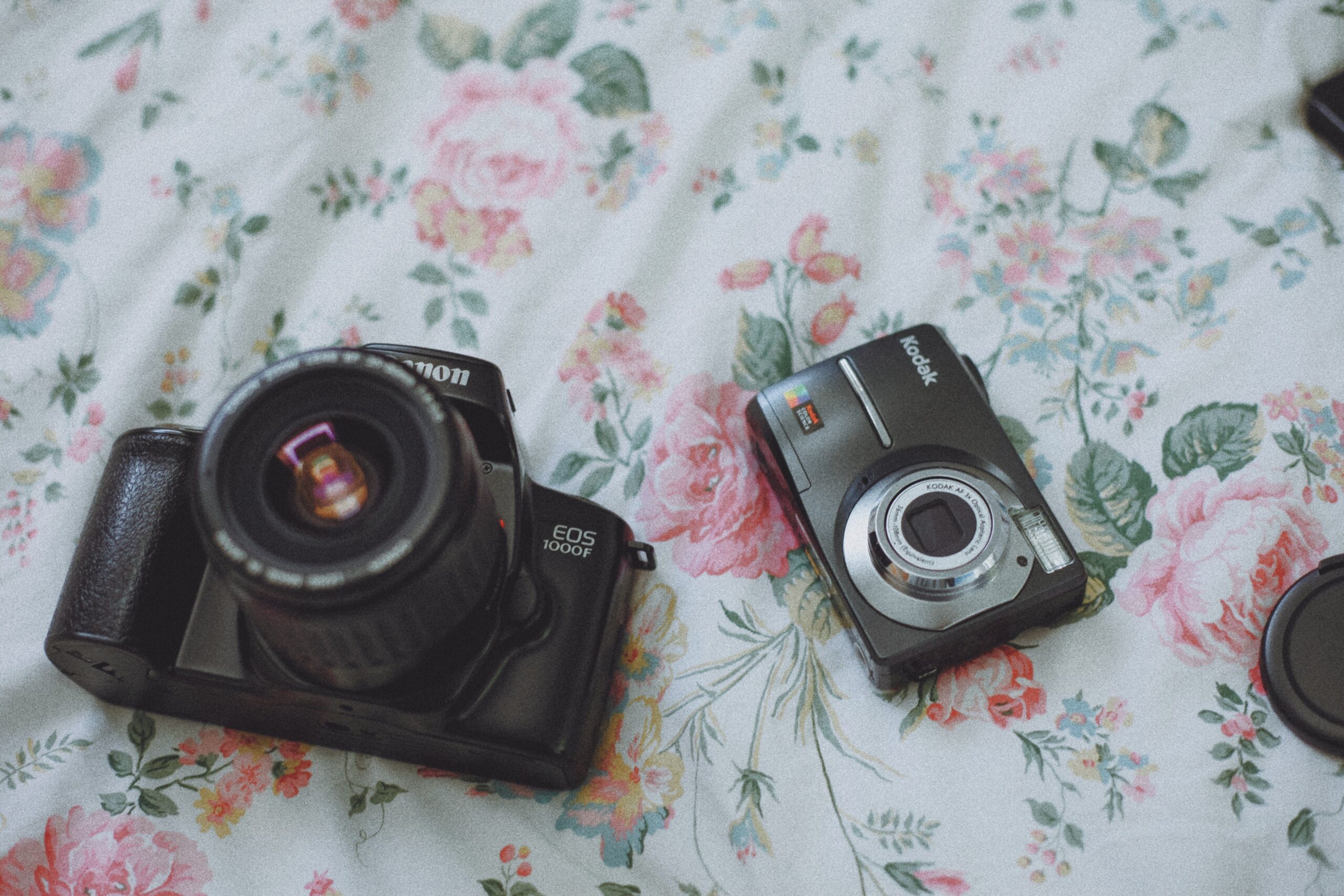 A DSLR and a compact camera are placed on a sheet of fabric with a floral pattern.